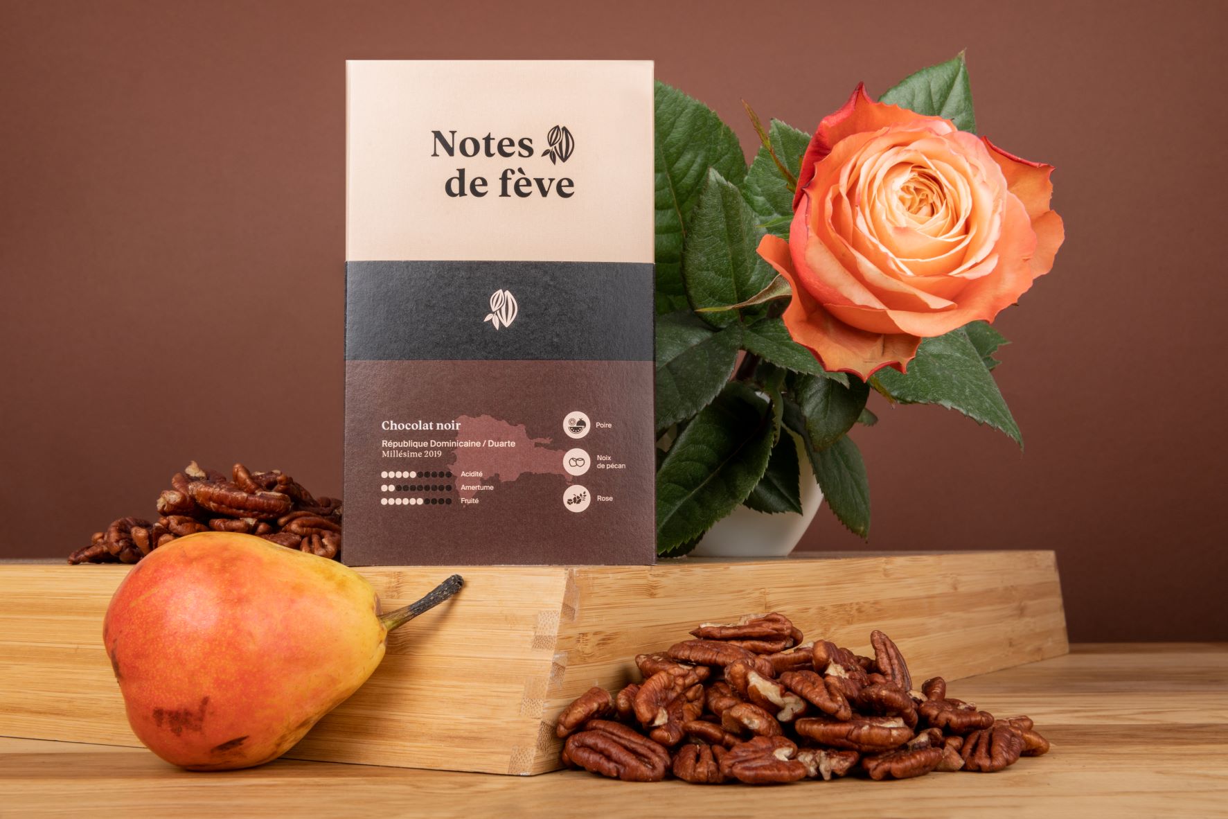 Une tablette de chocolat avec un emballage brun. Autour, on voit une rose, une poire et des noix de pécan, trois arômes que l'on retrouve naturellement dans la fève de cacao.