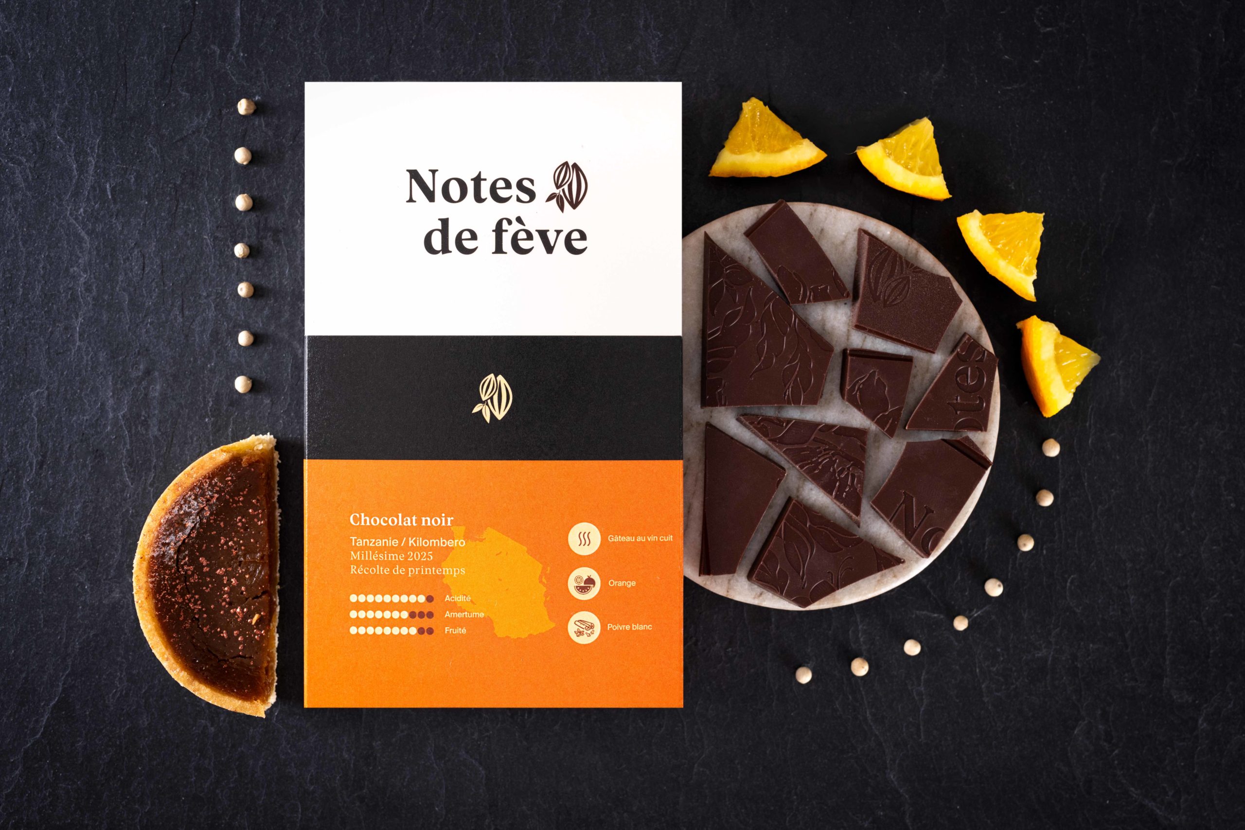 Une tablette de chocolat Bean to bar avec un emballage orange. Autour, du gâteau au vin cuit, de l'orange et du poivre blanc.