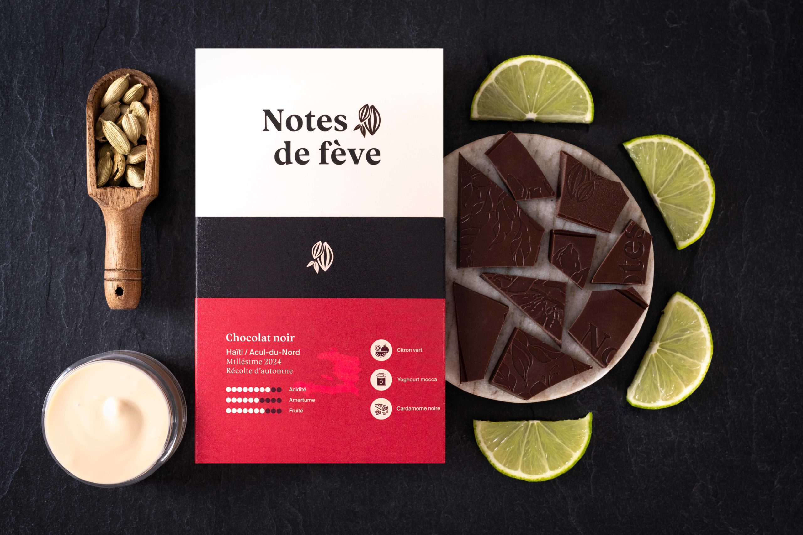 Une tablette de chocolat bean to bar avec un emballage rouge. Autour, du citron vert, du yogourt moccas et de la caradamome
