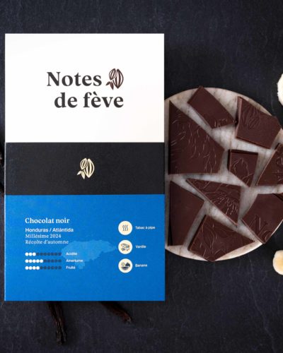 Une tablette de chocolat avec un emballage bleu. Autour, des bananes, des bâtons de vanille et du tabac