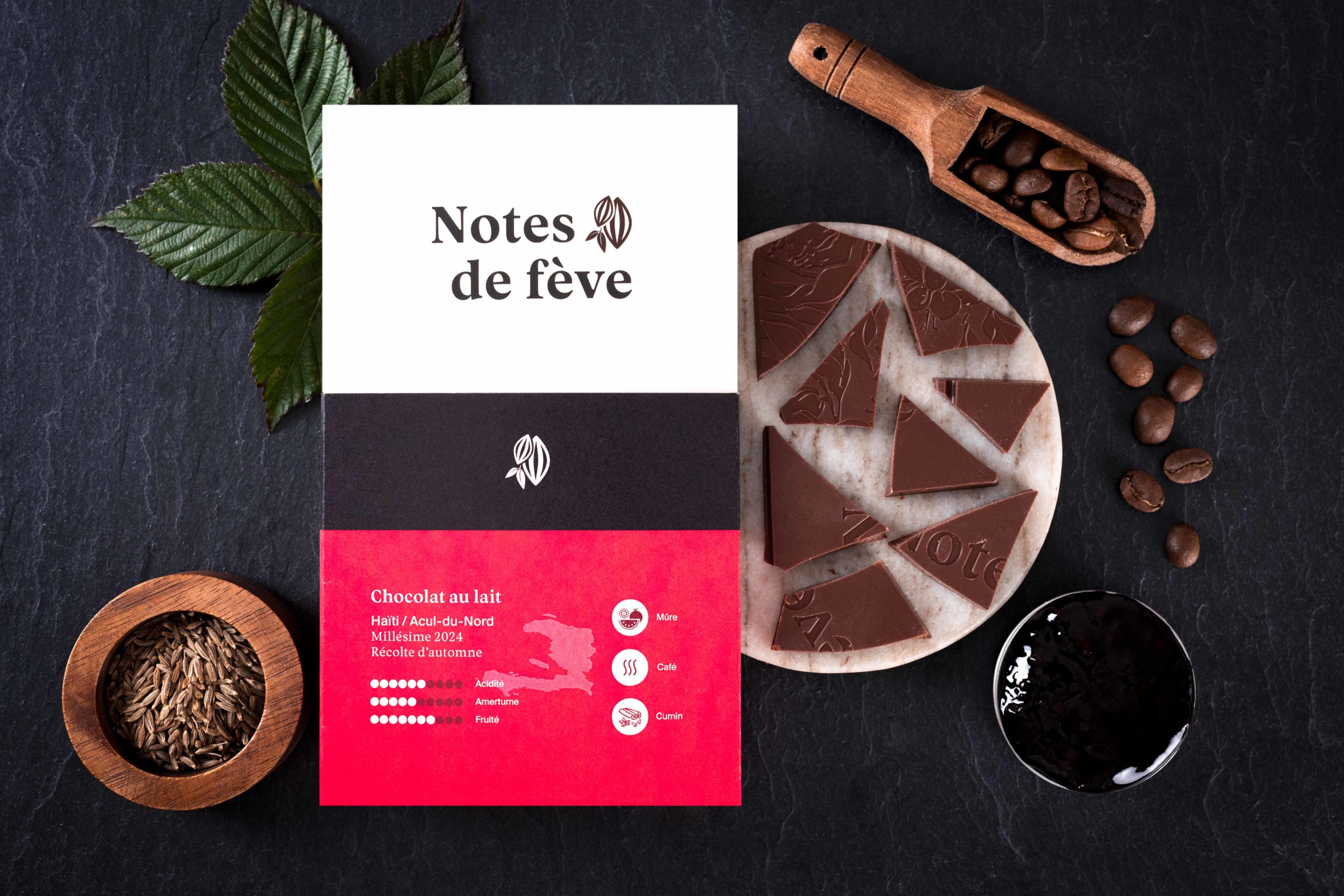 Une tablette de chocolat avec un emballage rose. Autour, on voit du cumin, des grains de café, ainsi que des feuilles de mûrier et de la confiture de mûres.
