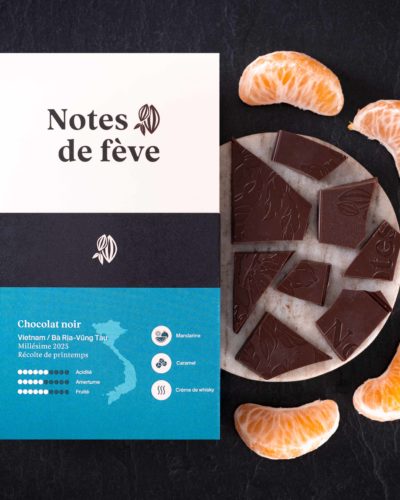 Une tablette de chocolat bean to bar avec un emballage bleu-vert. Autour, des mandarines, du caramel et de la crème de whisky