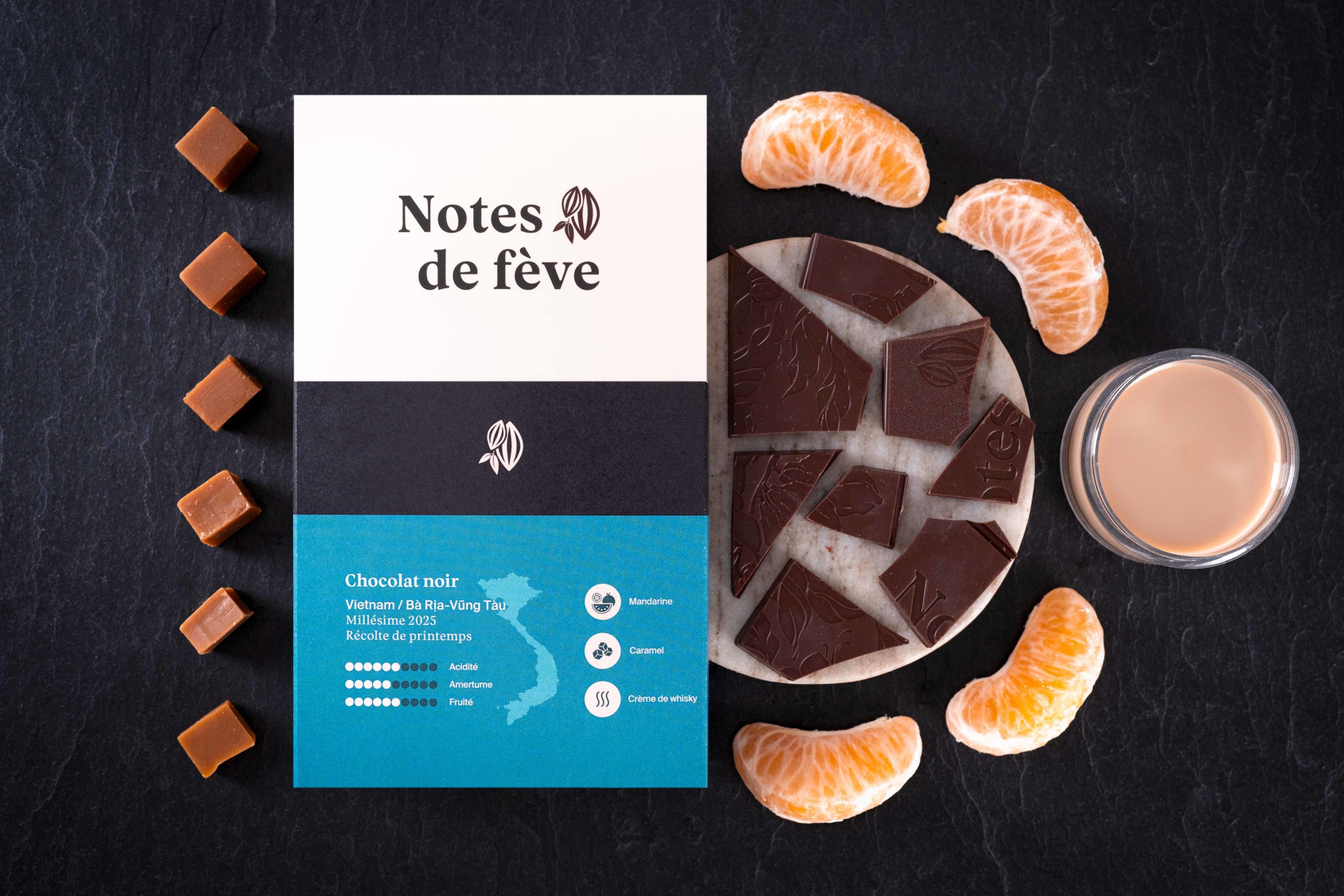 Une tablette de chocolat bean to bar avec un emballage bleu-vert. Autour, des mandarines, du caramel et de la crème de whisky