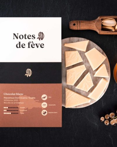 Une tablette de chocolat avec un emballage beige. Autour, du miel, des raisinets blancs et des cacahuètes