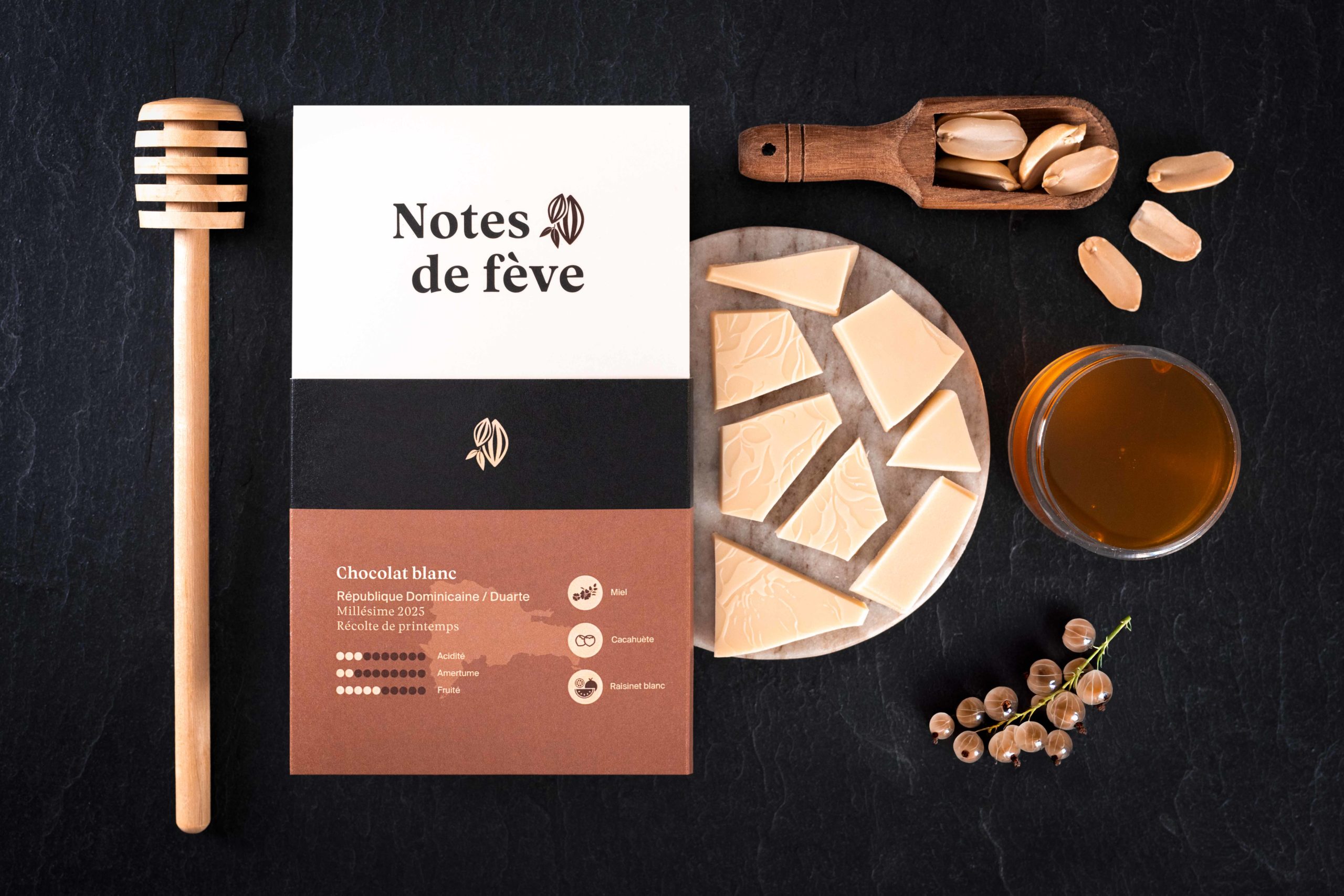 Une tablette de chocolat avec un emballage beige. Autour, du miel, des raisinets blancs et des cacahuètes