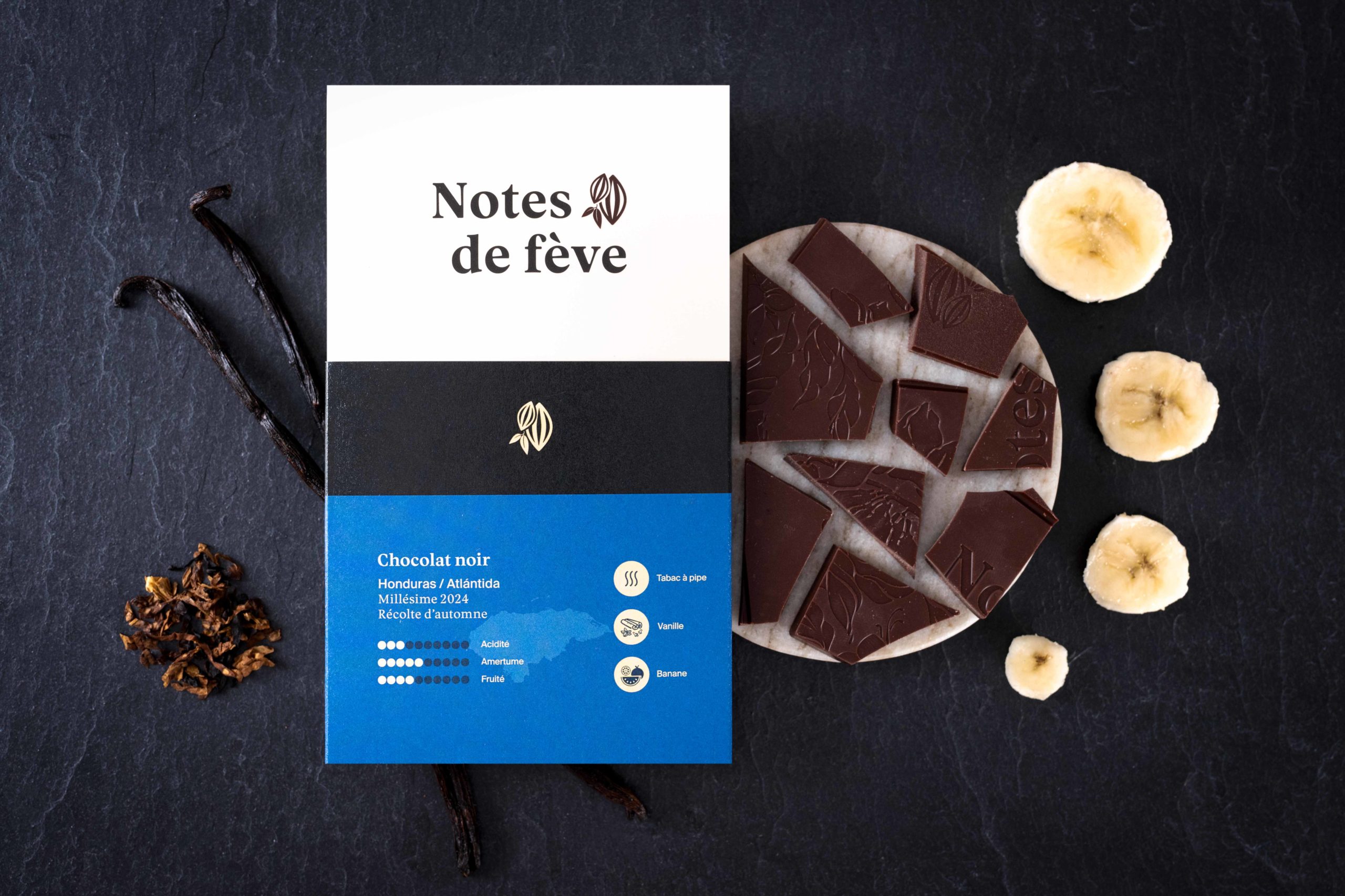 Une tablette de chocolat avec un emballage bleu. Autour, des bananes, des bâtons de vanille et du tabac