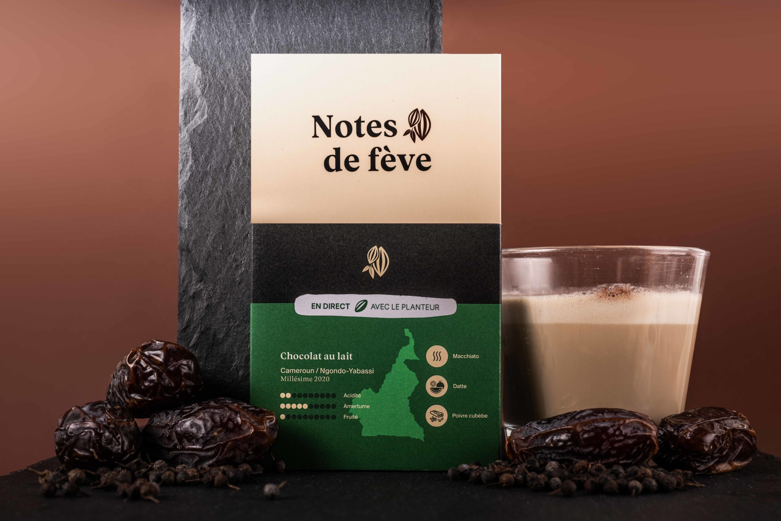 Une tablette de chocolat au lait à l'emballage vert, entourée par des dattes, du poivre, et un verre de Latte Macchiato