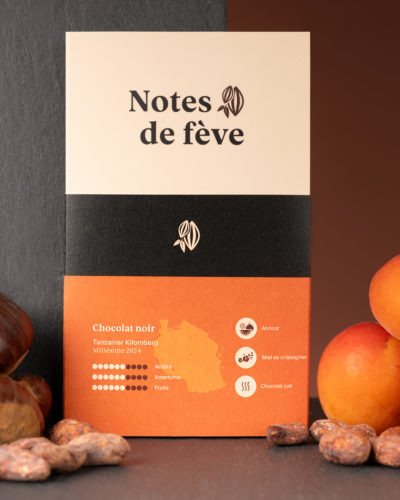 Une tablette de chocolat avec un emballage orange. Autour de la tablette, on voit des abricots, des fève de cacao et des châtaignes.